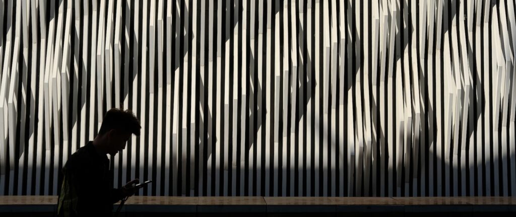 Fotografia em cores, mas dominada pelo preto e pelo branco. Em primeiro plano, um homem, na China, caminha olhando pro celular. Atrás dele, um painel de textura em ripas brancas finas sobre fundo preto.