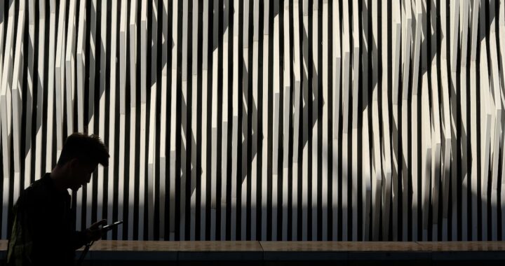 Fotografia em cores, mas dominada pelo preto e pelo branco. Em primeiro plano, um homem, na China, caminha olhando pro celular. Atrás dele, um painel de textura em ripas brancas finas sobre fundo preto.