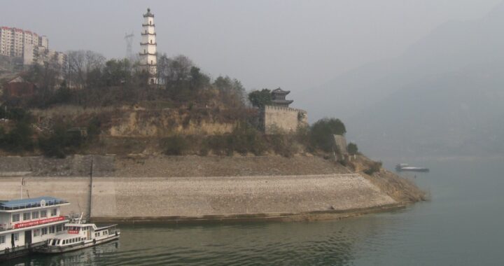 Fotografia em cores do rio Yangtze. Vê-se um barco na água e um trecho da margem se elevando, com prédios chineses tradicionais e modernos ao fundo. Há uma névoa ofuscando o fundo da paisagem.