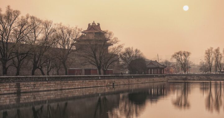 Imagem de cenário em Pequim com filtro amarelado. Vê-se um rio, as árvores secas e calçada à sua esquerda, um prédio tradicional ao fundo e o sol.
