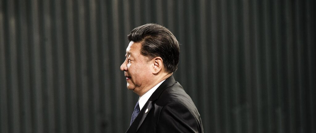 Fotografia em cores de Xi Jinping de perfil, vestindo um terno preto e camisa branca, sob fundo escuro ao ar livre.