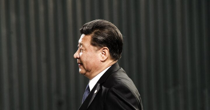Fotografia em cores de Xi Jinping de perfil, vestindo um terno preto e camisa branca, sob fundo escuro ao ar livre.