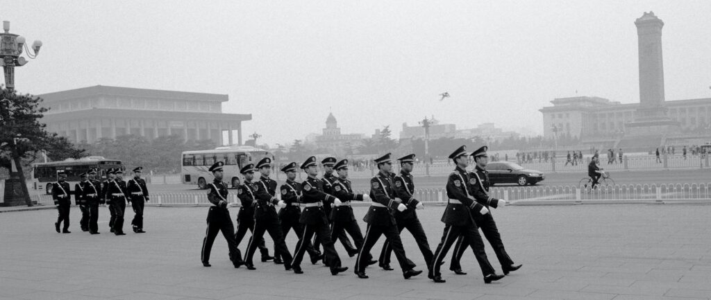 Fotografia em preto e branco, contemporânea, de soldados marchando uniformizados na Praça da Paz Celestial.