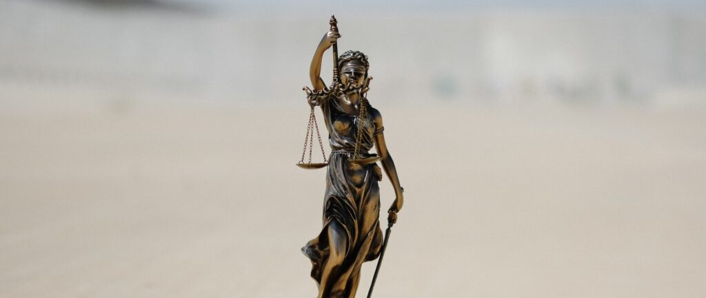 Fotografia em cores de estátua representando a justiça - uma mulher de olhos vendados segurando uma balança com dois pratos equilibrados.