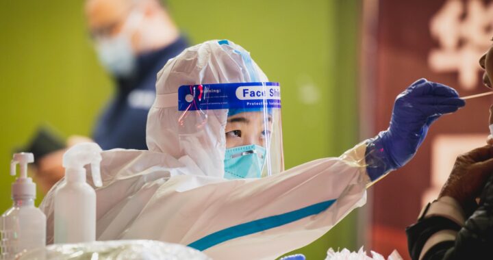 Fotografia em cores de trabalhadora da saúde chinesa em trajes de proteção bioquímica realizando coleta para exame de Covid-19 em pessoa fora d foco na foto. A foto foi tirada em Shenzhen.