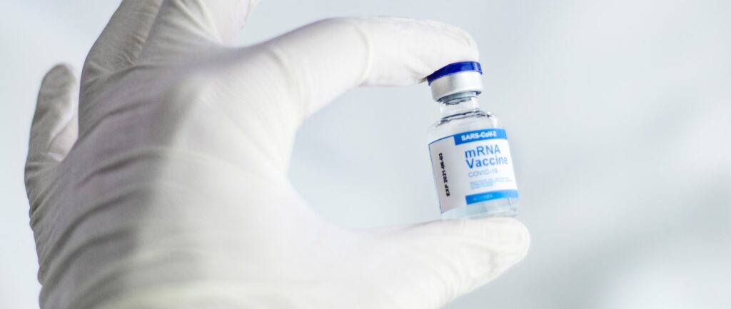 Fotografia em cores de mão em luva de látex, segurando um frasco de vacina mRNA contra Covid-19.