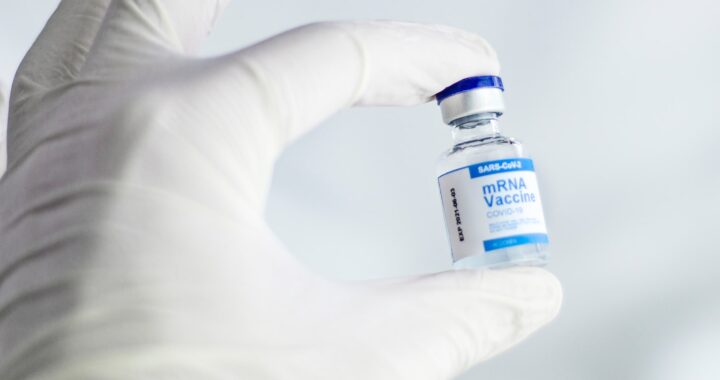 Fotografia em cores de mão em luva de látex, segurando um frasco de vacina mRNA contra Covid-19.