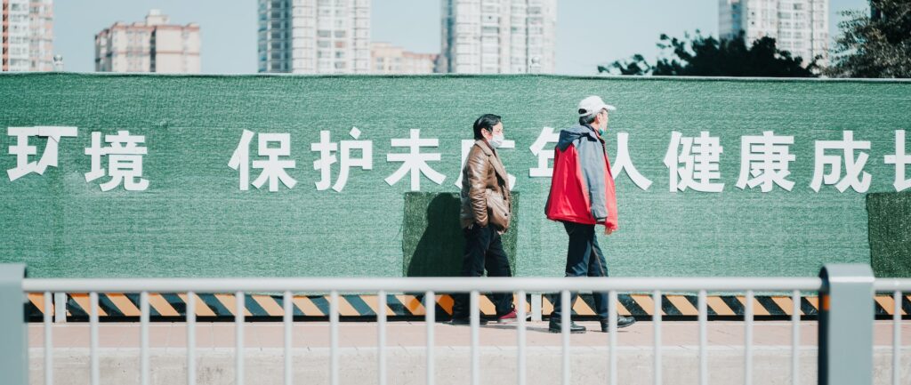 Fotografia em cores de casal idoso caminhando em dia de sol, um deles usa máscara e o outro está com a máscara no queixo. Atrás deles, um muro verde com palavras em chinês escritas em branco. Ao fundo, alguns prédios.