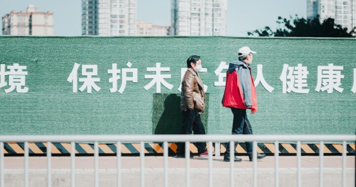 Fotografia em cores de casal idoso caminhando em dia de sol, um deles usa máscara e o outro está com a máscara no queixo. Atrás deles, um muro verde com palavras em chinês escritas em branco. Ao fundo, alguns prédios.