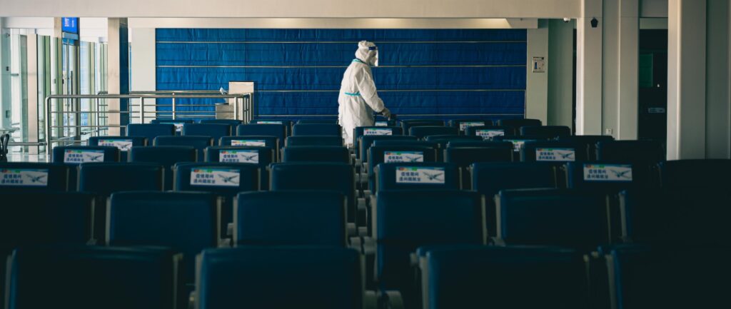 Uma pessoa em roupa de contenção branca, máscara e face-shield está numa sala escura, atrás de muitas cadeiras azuis e vazias.