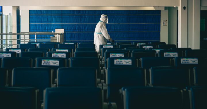 Uma pessoa em roupa de contenção branca, máscara e face-shield está numa sala escura, atrás de muitas cadeiras azuis e vazias.