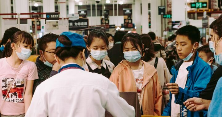 Fotografia em cores de um mercado chinês. Um atendente, de costas, chapeia carnes. Clientes estão ao seu redor, alguns de máscaras, outros com a máscara no queixo.
