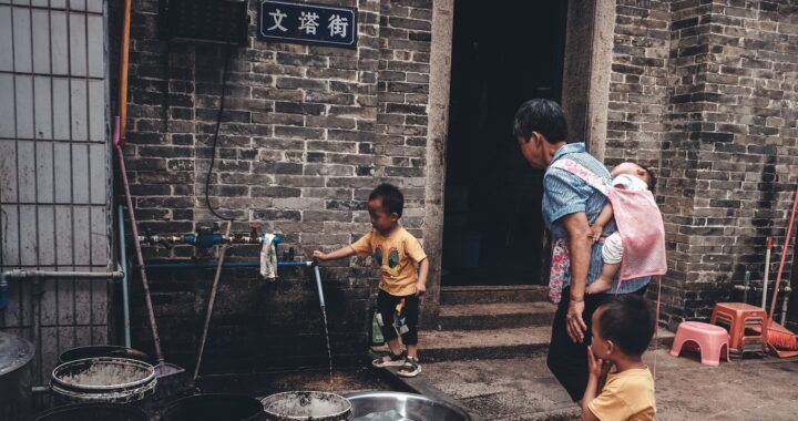 Foto de criança chinesa abrindo uma torneira de água do lado de fora de uma casa chinesa de tijolos cinzas. Com a criança há uma senhora, carregando um bebê no corpo e de mão dada com outro menino.