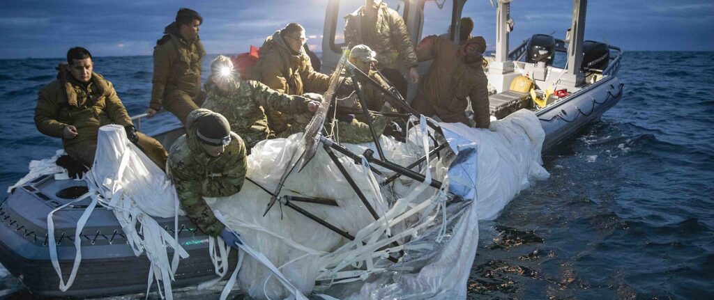 Fotografia em cores de marinha dos EUA resgatando o balão chinês no oceano. Pode-se ver marinheiros em um bote, no começo da noite, puxando o balão para dentro da embarcação.