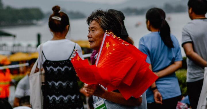 Fotografia em cores de idoso chinês carregando bandeiras da China. Ao fundo, pessoas observam um rio.