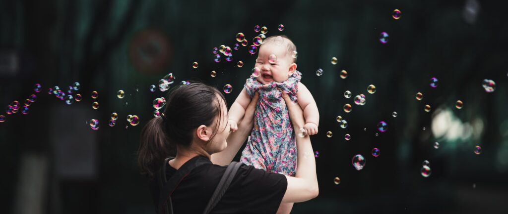 Fotografia em cores de bebê chinesa sendo erguida pela mãe. Ao fundo, árvores, e ao redor delas há bolhas de sabão.