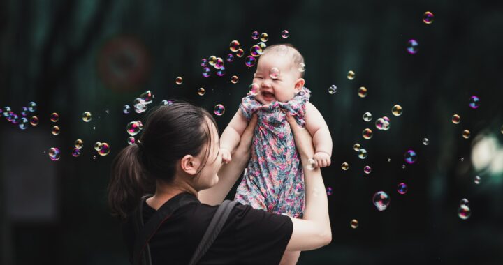 Fotografia em cores de bebê chinesa sendo erguida pela mãe. Ao fundo, árvores, e ao redor delas há bolhas de sabão.