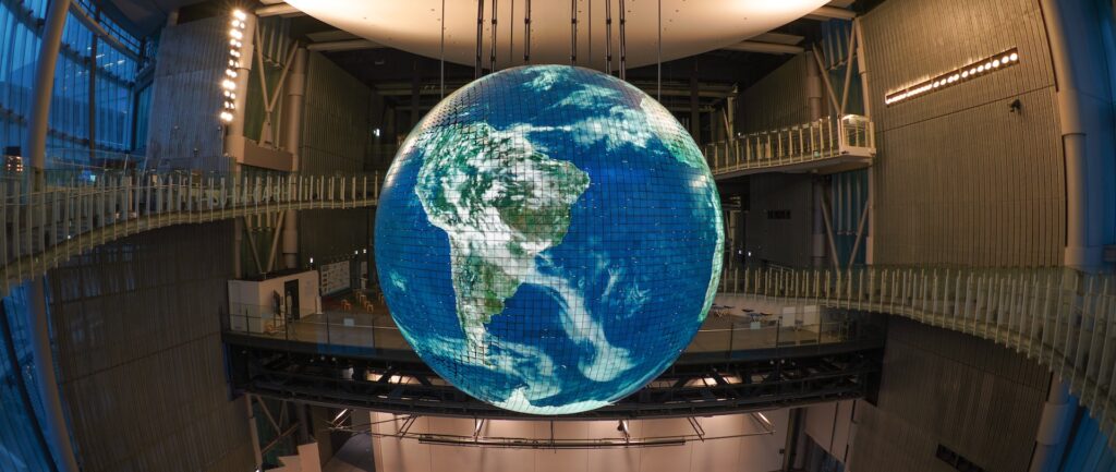 Fotografia em cores de globo iluminado, com a América Latina em primeiro plano. O globo é grande e pende do teto de um salão espaçoso.