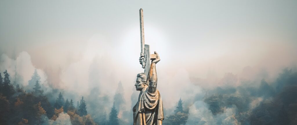 Fotografia em cores do Monumento à mãe pátria em Kiev, na Ucrânia. A estátua de um homem de braços erguidos, segurando um escudo e uma espada, está cercada de um nevoeiro. Ao fundo, árvores.