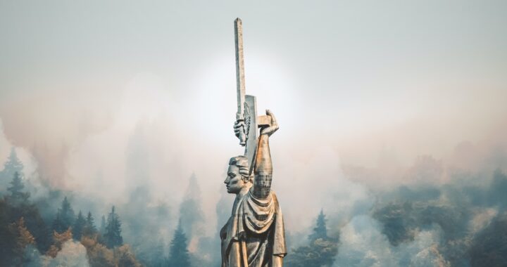 Fotografia em cores do Monumento à mãe pátria em Kiev, na Ucrânia. A estátua de um homem de braços erguidos, segurando um escudo e uma espada, está cercada de um nevoeiro. Ao fundo, árvores.