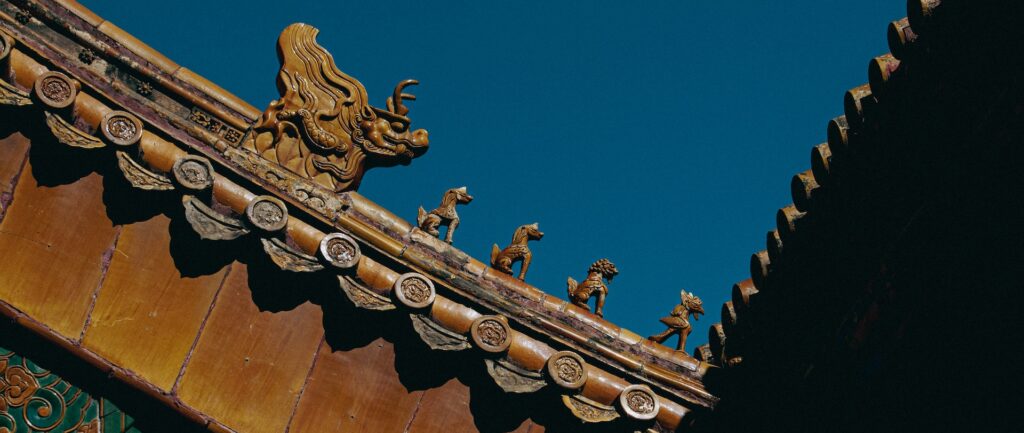 Fotografia em cores de detalhe de um telhado no Museu do Palácio em Pequim. Vê-se uma fileira de pequenas esculturas, sendo um dragão e, à sua frente, quatro criaturas menores.