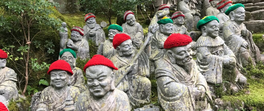 Fotografia em cores de estátuas religiosas de pedra em templo de Hiroshima. Algumas estátuas sorriem, outras têm expressão grave; algumas usam chapéus vermelhos, outras verdes.