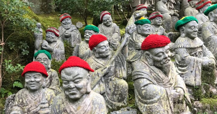 Fotografia em cores de estátuas religiosas de pedra em templo de Hiroshima. Algumas estátuas sorriem, outras têm expressão grave; algumas usam chapéus vermelhos, outras verdes.