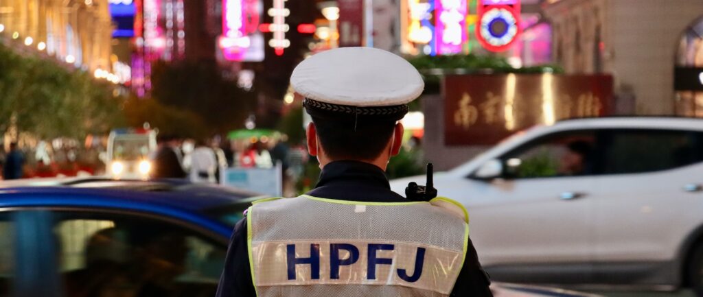 Fotografia em cores de rua em Shanghai. Em foco, um policial de costas, HPFJ escrito em seu colete. Ao fundo, carros em movimento e luminosos de neon.