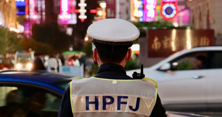 Fotografia em cores de rua em Shanghai. Em foco, um policial de costas, HPFJ escrito em seu colete. Ao fundo, carros em movimento e luminosos de neon.