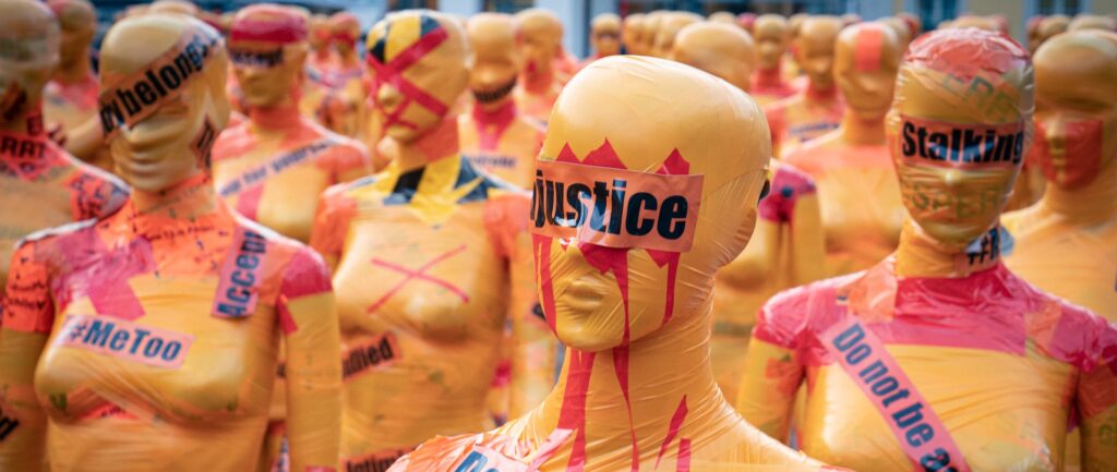 Imagem de protesto contra a violência de gênero. Manequins femininos envelopados com plástico amarelo, enrolados com fita plástica laranja com palavras-chave como #MeToo, stalking e justice, com adesivos vermelhos em forma de xis.