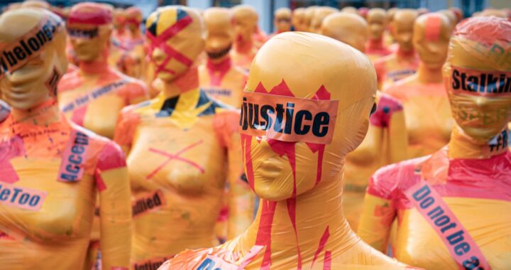 Imagem de protesto contra a violência de gênero. Manequins femininos envelopados com plástico amarelo, enrolados com fita plástica laranja com palavras-chave como #MeToo, stalking e justice, com adesivos vermelhos em forma de xis.