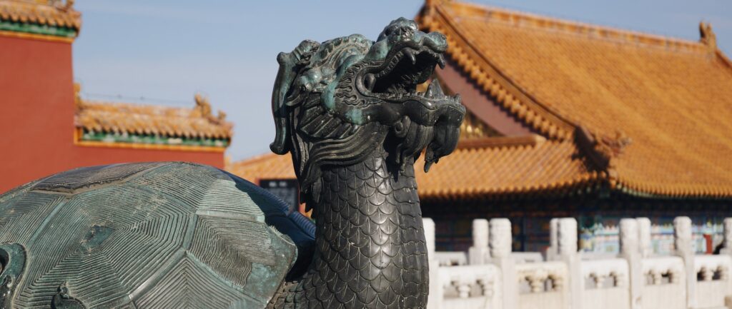 Fotografia em cores da estátua de tartaruga dragão na Cidade Proibida. A estátua de bronze esverdeado está em primeiro plano, com telhados laranjas e muletas brancas fora de foco ao fundo.