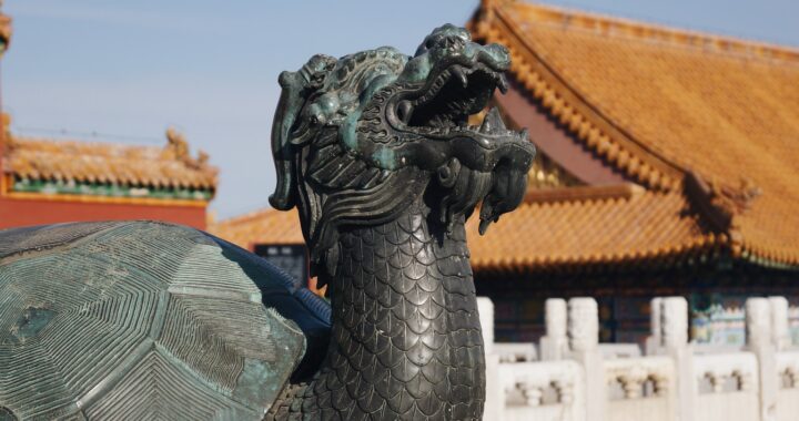 Fotografia em cores da estátua de tartaruga dragão na Cidade Proibida. A estátua de bronze esverdeado está em primeiro plano, com telhados laranjas e muletas brancas fora de foco ao fundo.