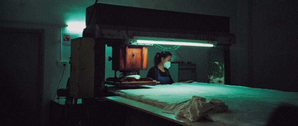 Fotografia em cores de trabalhadora em fábrica. Ao redor da mulher, é possível ver maquinário iluminado por uma luz branca-esverdeada. A mulher usa uma máscara facial.