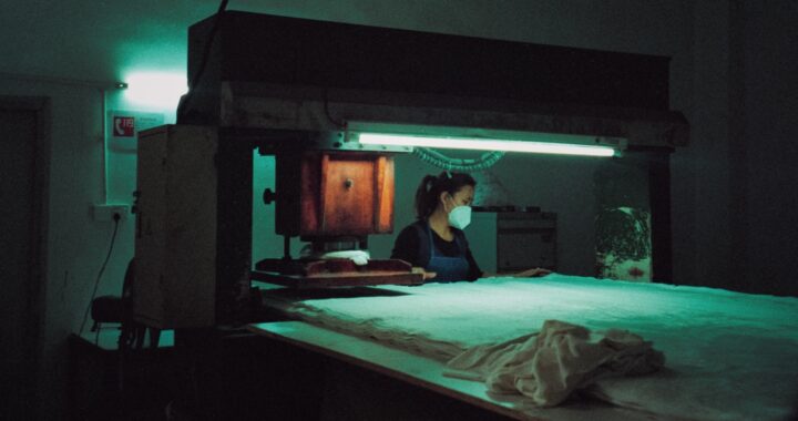 Fotografia em cores de trabalhadora em fábrica. Ao redor da mulher, é possível ver maquinário iluminado por uma luz branca-esverdeada. A mulher usa uma máscara facial.