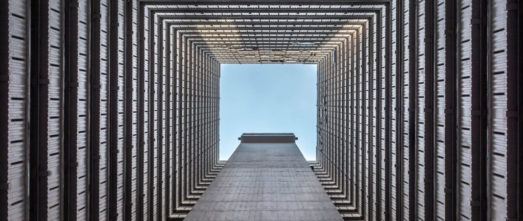 Fotografia em cores de céu azul visto a partir da base da área vazada de um prédio. O prédio é fechado dos quatro lados.