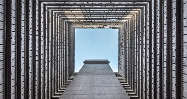 Fotografia em cores de céu azul visto a partir da base da área vazada de um prédio. O prédio é fechado dos quatro lados.