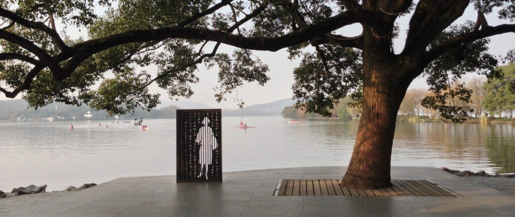 Fotografia em cores de parque à beira de um rio. Sob uma árvore, há uma instalação artística representando, em branco sobre material preto, um perfil feminino.