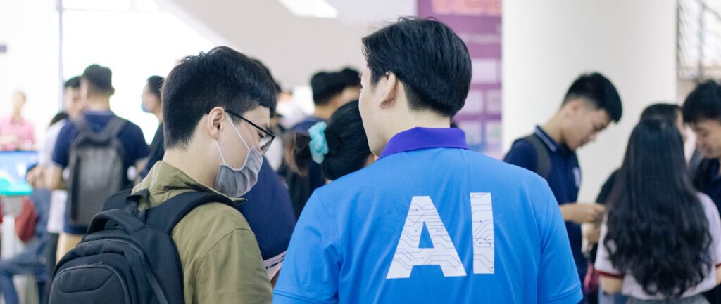 Foto em cores de um homem chinês, de costas, com AI escrito na camiseta azul. Ele fala com outro homem chinês, de máscara no rosto e mochila nas acostas. Ao fundo, diversos jovens conversam em grupos.