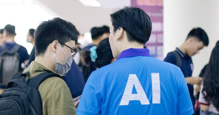 Foto em cores de um homem chinês, de costas, com AI escrito na camiseta azul. Ele fala com outro homem chinês, de máscara no rosto e mochila nas acostas. Ao fundo, diversos jovens conversam em grupos.
