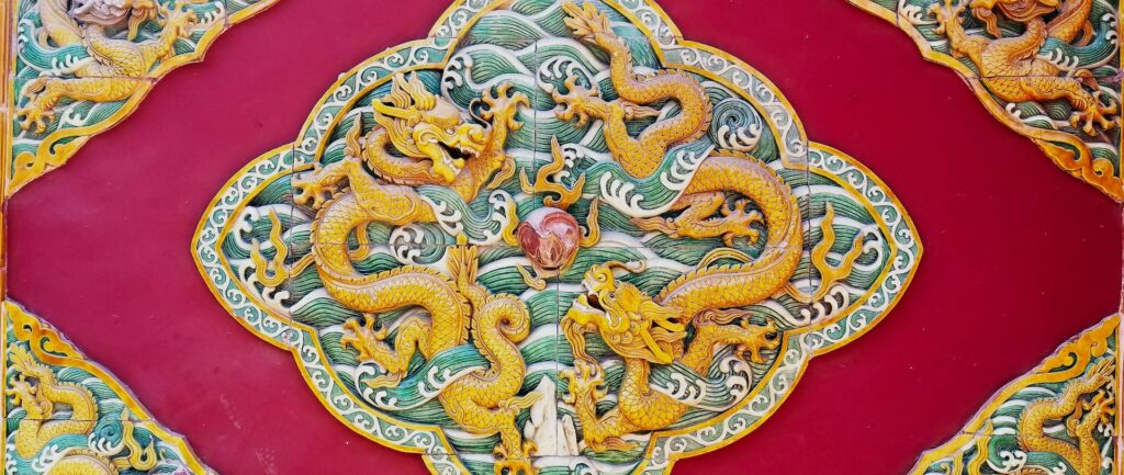 Fotografia em cores de detalhe da decoração em templo no Beihai Park, mostrando dois dragões dourados ao redor de uma pedra vermelha.