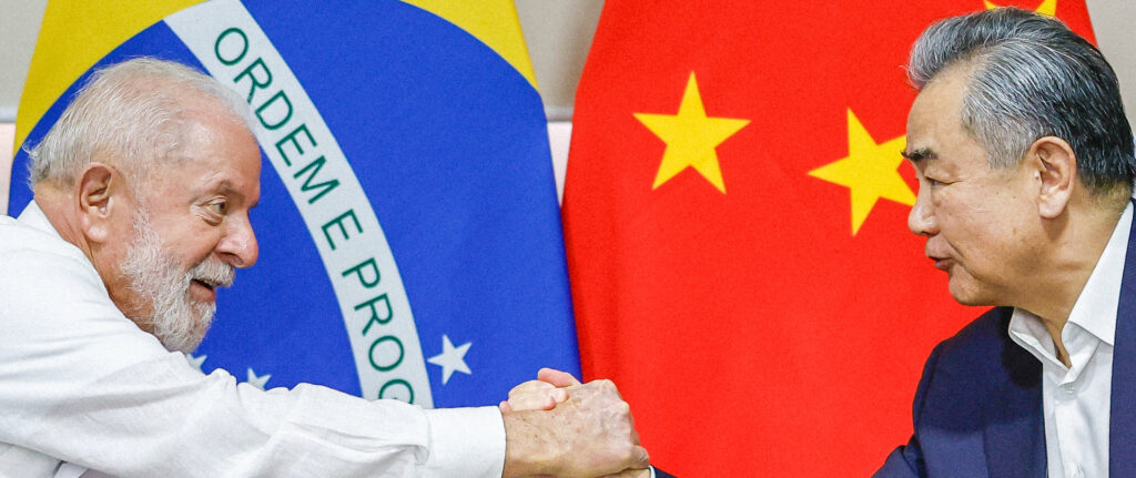 Imagem do presidente Lula vestindo camisa branca e apertando a mão do chanceler chinês Wang Yi, com terno azul marinho e camisa branca, sem gravata. Ao fundo estão bandeiras brasileira e chinesa. Copos de água abaixo deles