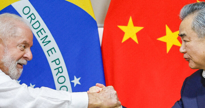 Imagem do presidente Lula vestindo camisa branca e apertando a mão do chanceler chinês Wang Yi, com terno azul marinho e camisa branca, sem gravata. Ao fundo estão bandeiras brasileira e chinesa. Copos de água abaixo deles