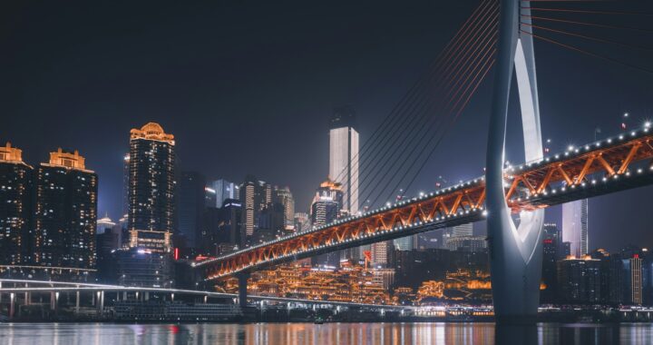 Fotografia em cores noturna de ponte sobre águas em Chongqing, na China. A ponte, iluminada, estaiada e com uma passagem aberta para os carros em seu pilar central, tem um efeito de alta-tecnologia. Ao fundo, arranha-céus iluminados.