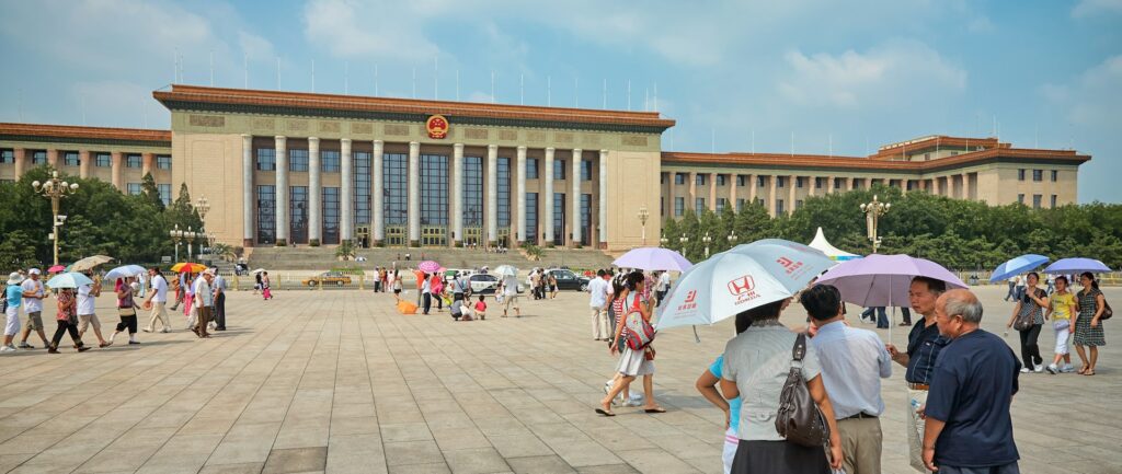 Fotografia em cores de pessoas com sombrinhas passeando pela Praça da Paz Celestial em dia de sol.