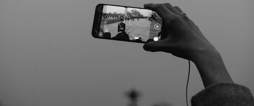 Fotografia em branco e preto de mão segurando um celular para tirar uma foto. No celular é possível ver militares em formação.