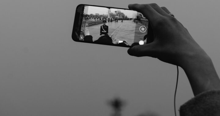 Fotografia em branco e preto de mão segurando um celular para tirar uma foto. No celular é possível ver militares em formação.