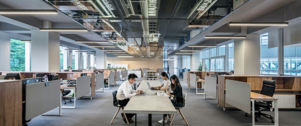Três jovens asiáticos sentados em uma longa mesa retangular no centro de um escritório. Eles vestem roupas formais de trabalhos em escritório e estão usando computadores e lendo revistas numa representação de trabalho em escritório. No teto há uma projeção digital de dados