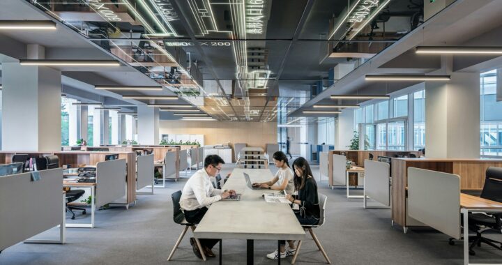 Três jovens asiáticos sentados em uma longa mesa retangular no centro de um escritório. Eles vestem roupas formais de trabalhos em escritório e estão usando computadores e lendo revistas numa representação de trabalho em escritório. No teto há uma projeção digital de dados