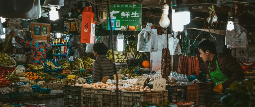 Fotografia em cores de barraca de mercado de rua em Shanghai. É possível ver dois vendedores e um cliente circulando entre caixotes de alimentos.